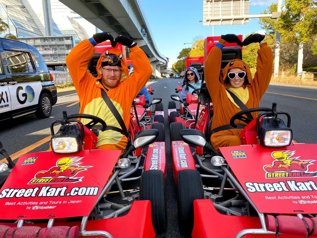 Two people in orange costumes in red go-karts on a city street, forming a heart with their arms as others follow behind