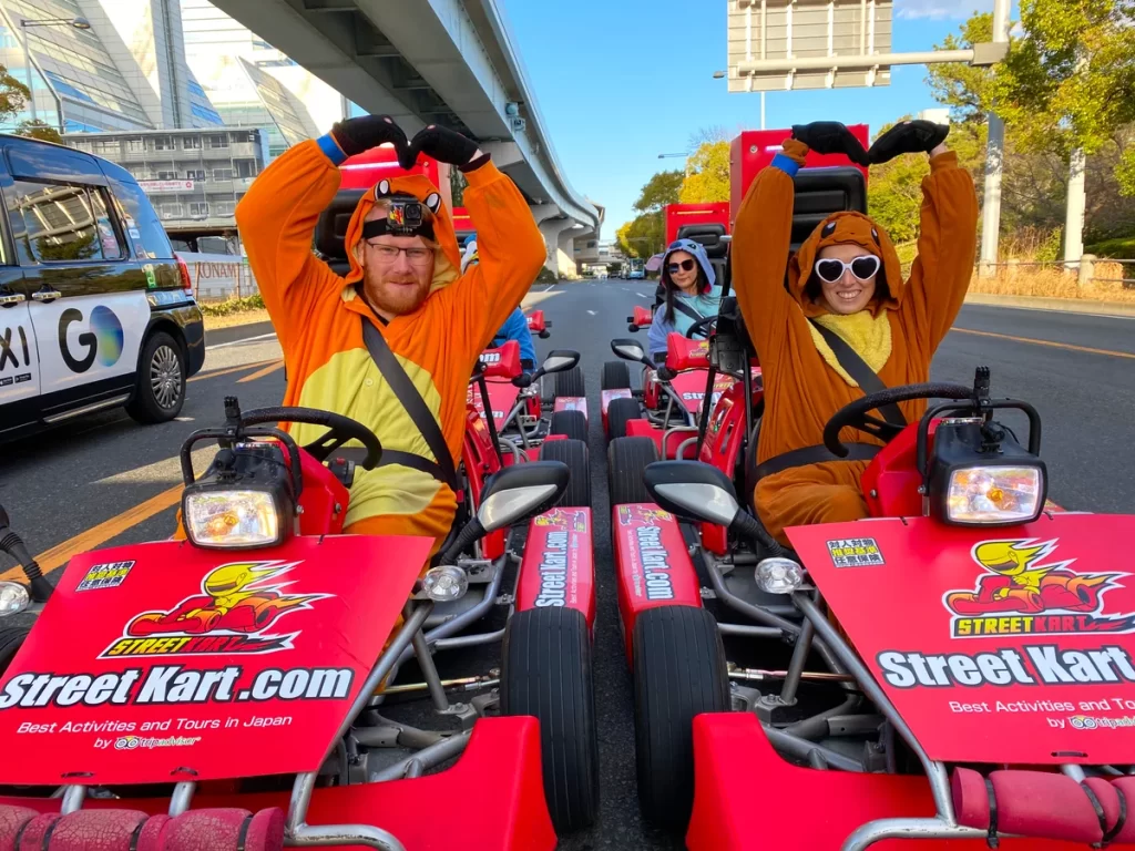 IMG_3605_converted - StreetKart Three people in orange animal onesies sit in bright red Street Kart go-karts on a city street, making heart shapes with their arms and smiling at the camera.