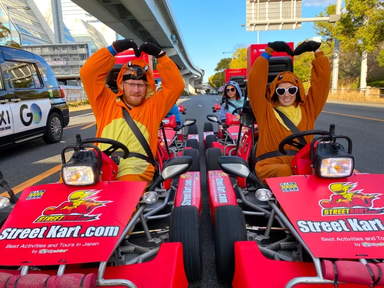 Three people in orange animal onesies sit in bright red Street Kart go-karts on a city street, making heart shapes with their arms and smiling at the camera.