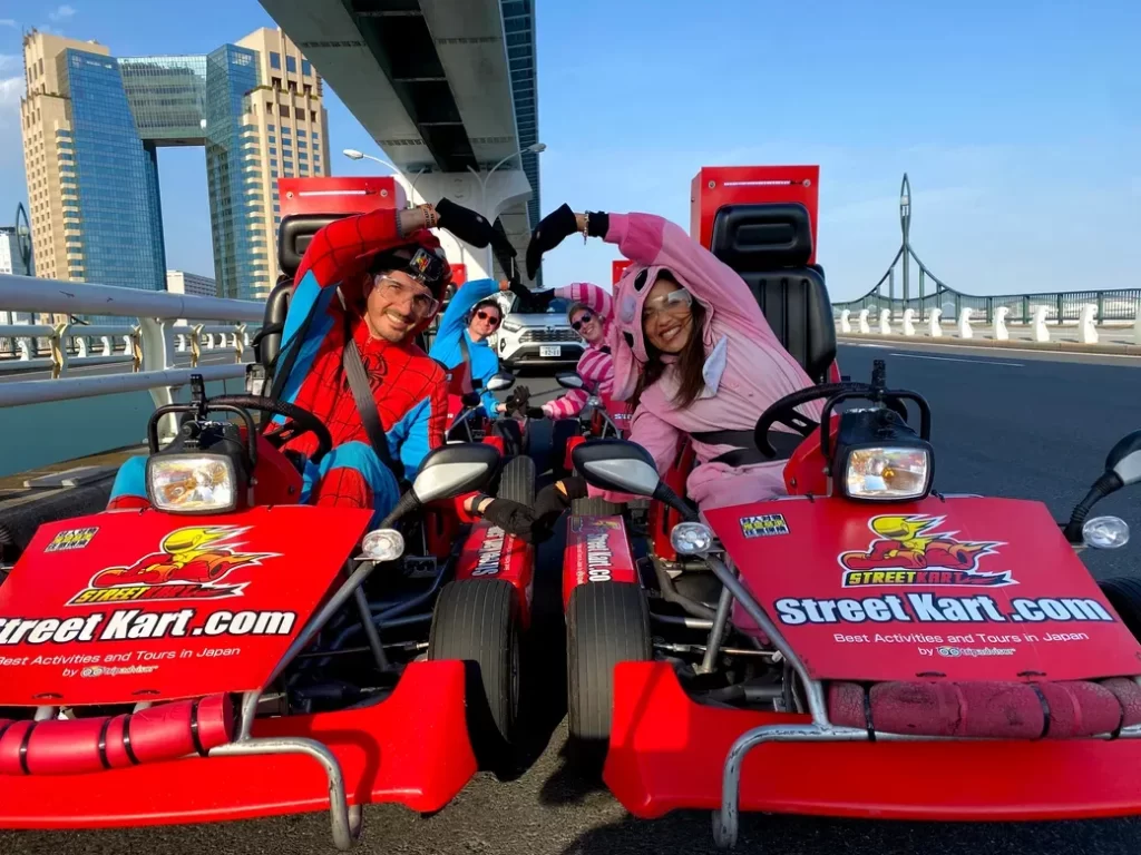 IMG_4112 - StreetKart Four friends in red go-karts on a sunny bridge, wearing Spider-Man and pink costumes, forming a heart with their arms.