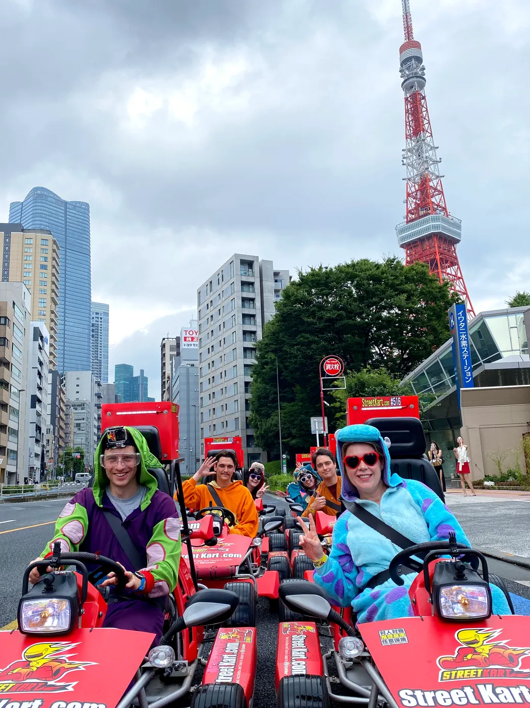 Group of people in colorful onesies driving red street-karts on a city street with Tokyo Tower in the background.